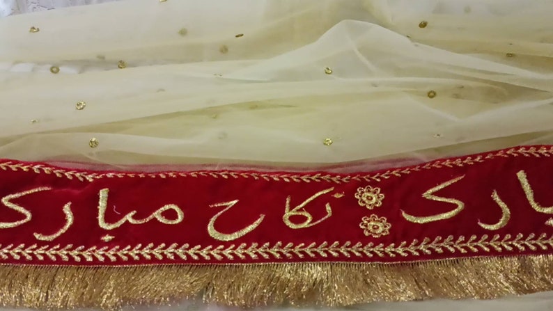 May include: A white sheer fabric with gold sequins and a red velvet trim with gold embroidery that reads "Ladi ki Maa"