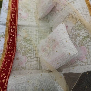 May include: A white bedspread with a red and gold trim. The bedspread is decorated with a delicate floral pattern and small gold beads. There are two white pillows on the bed, one with a floral pattern and the other with a plain white surface.