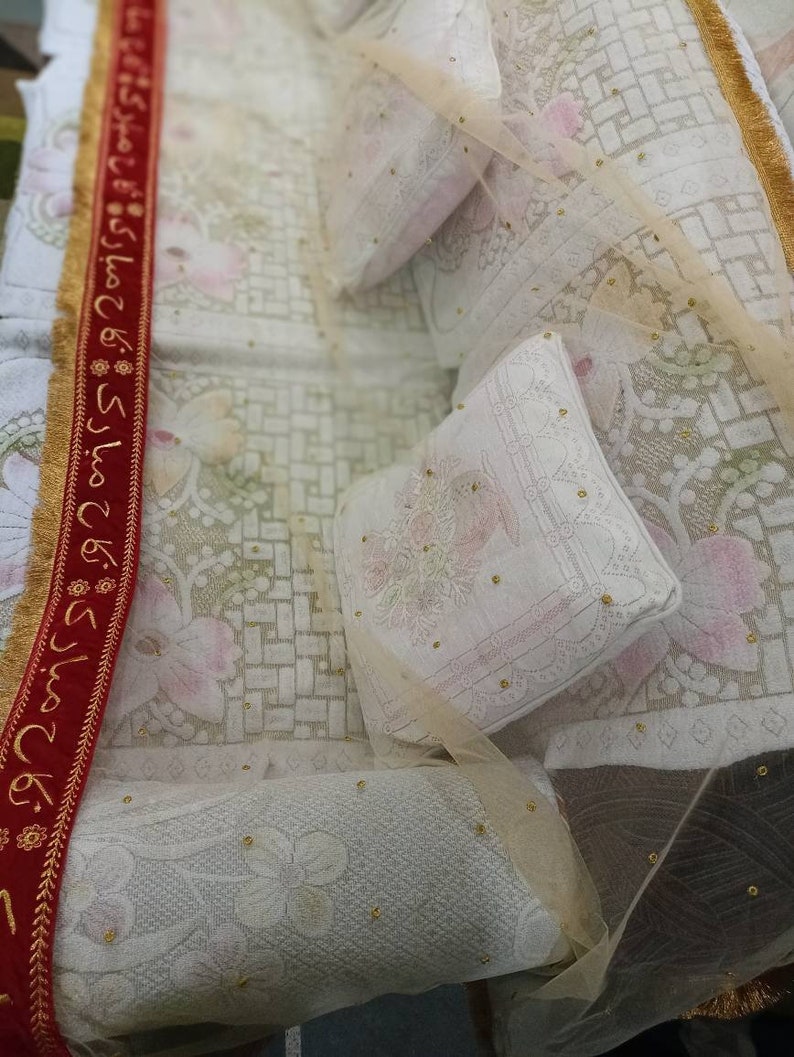 May include: A white bedspread with a red and gold trim. The bedspread is decorated with a delicate floral pattern and small gold beads. There are two white pillows on the bed, one with a floral pattern and the other with a plain white surface.