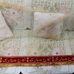 May include: A white couch with a floral pattern and gold accents. Two white pillows with gold accents are on the couch. A red ribbon with gold trim and the text "بارک اللہ کا مبارک ہو" is at the bottom of the image.