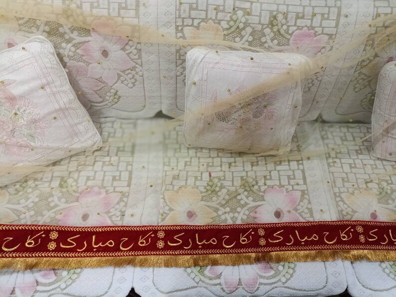 May include: A white couch with a floral pattern and gold accents. Two white pillows with gold accents are on the couch. A red ribbon with gold trim and the text "بارک اللہ کا مبارک ہو" is at the bottom of the image.