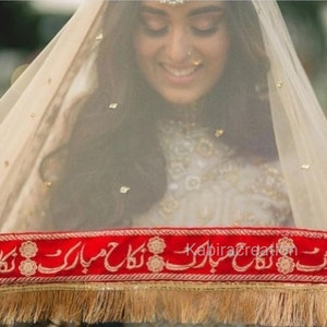 May include: A white veil with gold embellishments and a red embroidered border with the text "Kabira Creation" in Urdu script. The veil is worn by a person with long dark hair and a white dress.