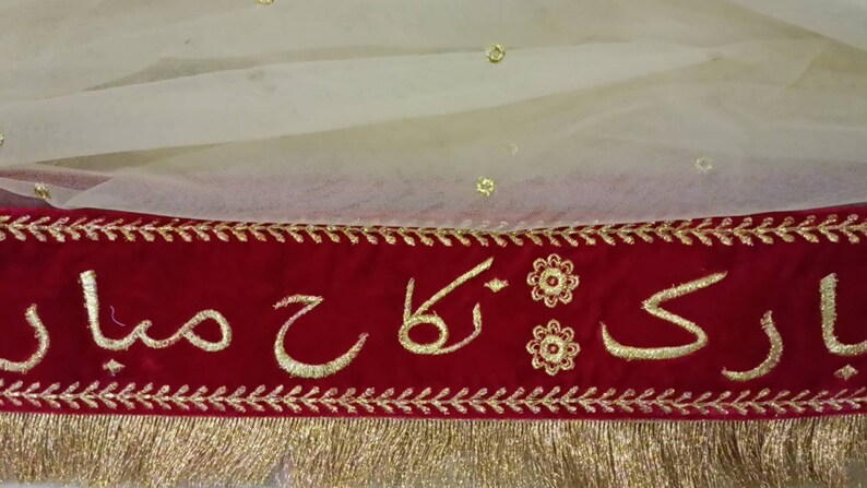 May include: A red velvet banner with gold embroidery featuring the words "Bari Ka Sajda" in Arabic script. The banner has a gold fringe trim and is decorated with small gold flowers.