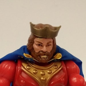 Puede incluir: Una figura de acción roja y dorada de un rey con una capa azul y una corona dorada. La figura está de pie sobre una base gris con un símbolo en ella. Un anillo dorado está en primer plano.