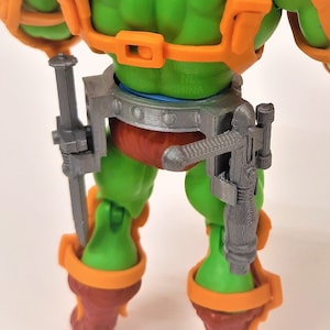 May include: A green action figure with orange armour, boots, and a blue helmet. The figure has a muscular physique and is equipped with two grey weapons attached to its waist. The figure is a collectible toy.