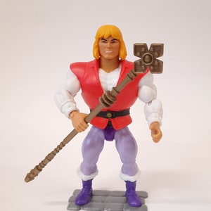 Puede incluir: Figura de acción vintage de un personaje musculoso con cabello rubio, que viste un chaleco rojo, mangas blancas y pantalones morados. La figura sostiene un bastón marrón con una parte superior en forma de cruz y está sobre una plataforma gris.
