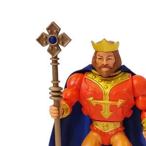 Puede incluir: Figura de acción vintage de un rey, vestido con armadura roja y dorada, una capa azul y botas. La figura tiene una corona dorada y sostiene un bastón con una cruz en la parte superior. Es un juguete coleccionable.