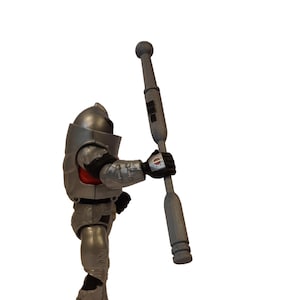 May include: A silver action figure in a futuristic armored suit, holding a gray staff. The figure has a helmet and red accents. The staff is cylindrical with a rounded top and bottom.
