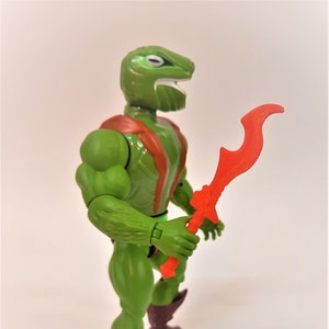 May include: Green action figure with a reptilian head and a red sword. The figure is wearing a brown and orange outfit.
