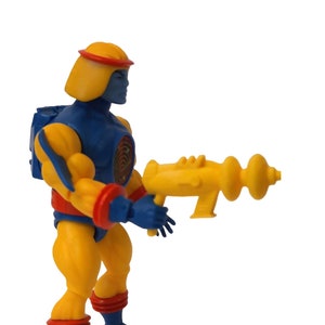 Puede incluir: Una figura de acción azul y amarilla con un casco rojo y amarillo y una pistola amarilla. La figura sostiene la pistola en su mano derecha.