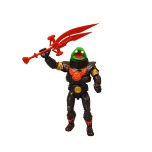 May include: A black action figure of a monster-like character with a green head and red mouth, holding a red weapon. The figure has gold accents and a red emblem on its chest. The toy is posed against a white background.