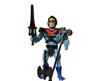 May include: A blue action figure with a yellow skull head and a black hood. The figure wears a black chest harness with a red bat emblem, black loincloth, and blue boots. It holds a black sword in one hand and a black staff in the other, with a black double-bladed axe standing at its feet.