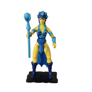 Puede incluir: Figura de acción vintage de un personaje femenino con un atuendo azul y amarillo, sosteniendo un bastón azul. La figura tiene un tocado y botas azules, con una capa azul. La figura está sobre una base negra.