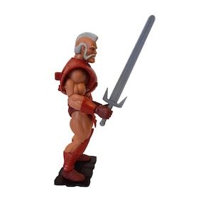 Puede incluir: Una figura de acción roja y bronceada de un hombre musculoso con barba y pelo blanco, sosteniendo una espada gris. La figura lleva una armadura y botas rojas. La figura está sobre una base negra.