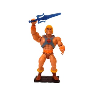 May include: A He-Man action figure with a blue sword held high. The figure has blonde hair, orange shorts, and a blue and orange chest piece. The figure stands on a brown base.