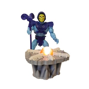 Peut inclure: Figurine de Skeletor avec un bâton, vêtu de bleu et de violet. Il se tient sur une plateforme grise avec un globe lumineux, décorée de structures en forme d'os. Une figurine de collection.
