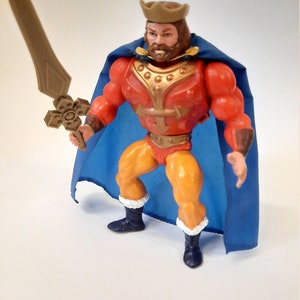Puede incluir: Una figura de acción vintage de un rey, vestido con armadura roja y dorada, una capa azul y una corona marrón. La figura sostiene una espada de color canela. La figura tiene piernas amarillas y botas negras con ribete blanco. La figura posa sobre un fondo blanco.