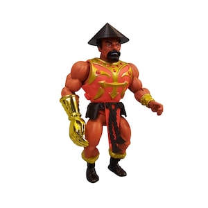 Op de afbeelding: Een gespierde actiefiguur met een gebruinde huid, een zwarte baard en een zwarte kegelvormige hoed. Het figuur draagt een rood borstpantser met gouden randen, een gouden handschoen aan de rechterarm, een gouden polsband links, zwarte laarzen en een zwarte lendendoek.