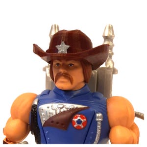 Könnte beinhalten: Ein brauner Cowboyhut mit einem silbernen Stern vorne, getragen von einer Figur mit einem braunen Schnurrbart und langen braunen Haaren. Die Figur trägt ein blaues Hemd mit einem silbernen und braunen Design.