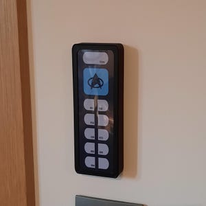 Op de afbeelding: Een rechthoekig zwart wandpaneel met een verticale opstelling van witte knoppen met zwarte tekst. De bovenste knop toont een blauw vierkant met een zwart Starfleet-insigne. Andere knoppen bevatten numerieke codes, waaronder 2308, 861, 150, 034, 320, 672, 880, 208 en 894.