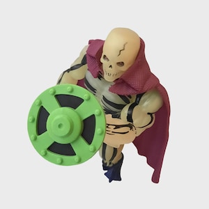 May include: A skeletal action figure with a light tan skull and muscular arms. It wears a dark pink cape and holds a green shield with a black center. The figure is posed as if in motion, with a detailed skeleton design.