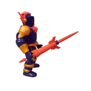 May include: A plastic action figure of a knight in dark purple armor with gold accents on the shoulders, arms, and legs. The figure wears a helmet with orange fin-like details and holds a long, pointed orange lance with a small fin guard.