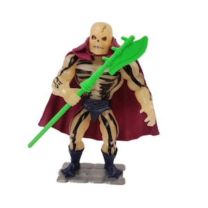 May include: A vintage action figure of a skeletal warrior with a green weapon and a burgundy cape. The figure has black stripes and tattoos on its arms. It stands on a gray, stone-like base.