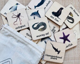May include: Wooden flashcards featuring sea creatures like a penguin, octopus, and hammerhead shark. Each card has a watercolor illustration and the creature's name. A canvas bag with the brand name "skills Toys" is visible.
