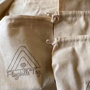 May include: A stack of natural cotton muslin drawstring bags with a logo that reads "Playskills Toys" printed on the front of one of the bags.