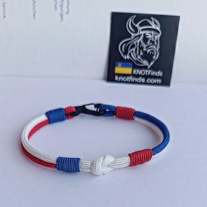 May include: A white, blue, and red paracord bracelet with a black clasp. The bracelet has a white knot in the center. The bracelet is made by KnotFinds.