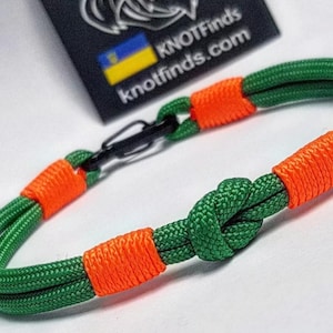 Puede incluir: Pulsera de cuerda verde y naranja con diseño de nudo. La pulsera tiene un cierre negro y detalles en naranja. La marca "KNOTFinds" es visible en una etiqueta negra con una bandera ucraniana.