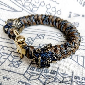 May include: A brown and blue paracord bracelet with a brass clasp and a detailed metal skull charm.