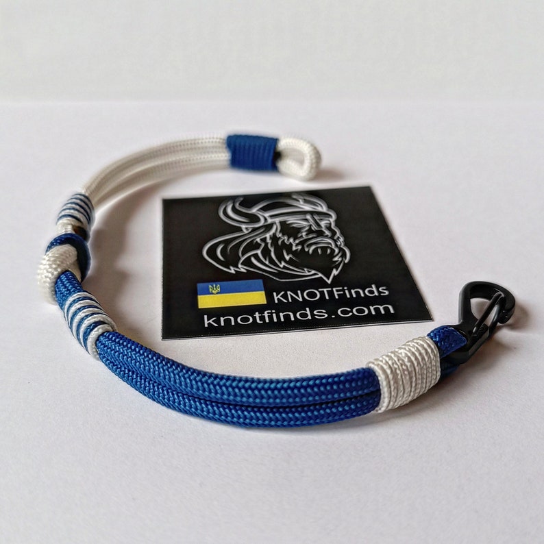 May include: A blue and white paracord bracelet with a black carabiner clip. The bracelet has a knot pattern and the words "KNOTFINDS knotfinds.com" are visible on a sticker in the background.