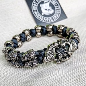 Könnte beinhalten: Ein schwarzes Paracord-Armband mit silbernen Totenkopfperlen und einem Totenkopfverschluss. Das Armband wird von Knot Viking Aggressive Bracelets hergestellt.