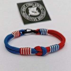Puede incluir: Una pulsera roja y azul con rayas blancas. La pulsera está hecha de un cordón tejido y tiene un cierre negro. La pulsera tiene un nudo en el centro y está decorada con rayas blancas. La pulsera está sobre una superficie blanca.