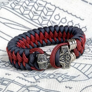 May include: A black and red paracord bracelet with a silver Mjolnir charm. The bracelet is braided and has a silver bead on the side.