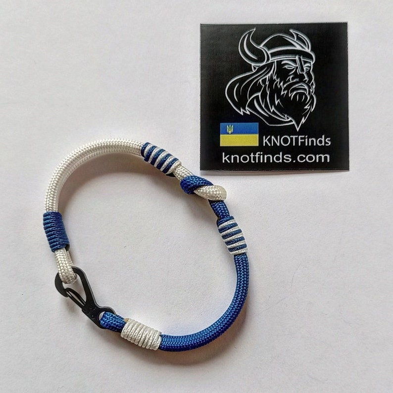 May include: A blue and white paracord bracelet with a silver clasp. The bracelet is tied in a knot pattern. The image also includes a black and white logo with the text "KNOTFinds knotfinds.com" and a Ukrainian flag.