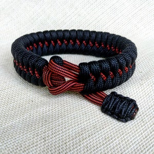 May include: Black and red paracord bracelet with a knot design. The bracelet is made of two strands of paracord, one black and one red, that are woven together to create a unique pattern.