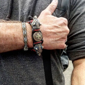 May include: A black paracord bracelet with a brass wolf head charm on each end and a brass cross charm in the center. The bracelet is worn on a person's wrist.