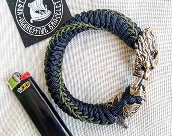 Puede incluir: Un brazalete de paracord negro y verde con un dije de cabeza de lobo de latón. El brazalete está hecho con un patrón de nudos y tiene un esquema de color negro y verde. El dije es una cabeza de lobo detallada con una superficie texturizada. El brazalete está sobre un fondo de tela blanca.