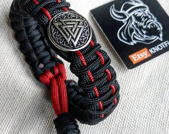 Op de afbeelding: Zwart en rood paracord armband met een zilveren metalen hanger met een Keltisch knoopontwerp. De hanger is rond en heeft een reliëf. Het armband is aan het ene uiteinde met een knoop vastgemaakt en heeft aan het andere uiteinde een metalen sluiting.