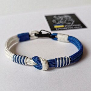 May include: A blue and white paracord bracelet with a black clasp. The bracelet has a knot in the center and is made of two strands of cord.