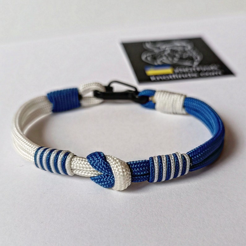 May include: A blue and white paracord bracelet with a black clasp. The bracelet has a knot in the center and is made of two strands of cord.