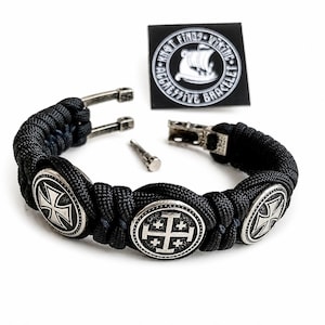 May include: Black braided bracelet with silver oval embellishments, each displaying a cross motif. The bracelet includes a metal clasp and a small pin. A black card with white text and a Viking ship logo is also visible.