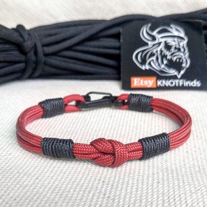 May include: Red and black paracord bracelet with a black metal clasp and a knot in the center. The bracelet is on a white fabric background. Etsy KNOTFINDS is visible on a black tag.