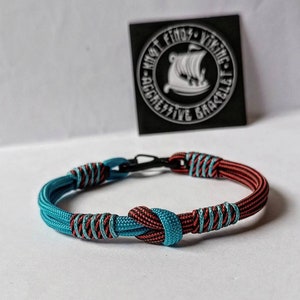 Op de afbeelding: Een blauw en rood paracord armband met een zwarte metalen sluiting. De armband is geknoopt met een knoop in het midden. De armband is gelabeld "What Finds Viking Aggressive Bracelet".