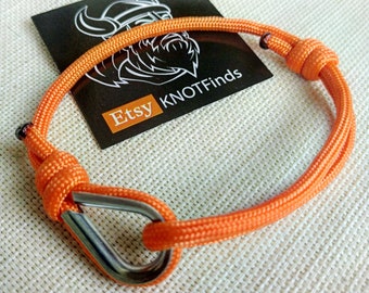 May include: Orange paracord bracelet with a silver carabiner clasp. The bracelet is tied with a knot on one end and has a tag that says "Etsy KNOTFinds".