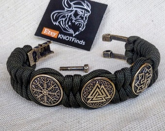 Op de afbeelding: Een groene paracord armband met drie metalen bedels met Noorse symbolen. De bedels zijn een Vegvisir, een Valknut en een Triskele. De armband heeft een metalen sluiting en is verstelbaar.