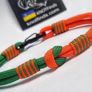 Op de afbeelding: Close-up van een groen en oranje touw armband. De armband heeft een knoopontwerp met oranje en groene accenten. Een zwarte sluiting is zichtbaar. De merknaam "knotfinds.com" staat op een zwart label met een Oekraïense vlag.