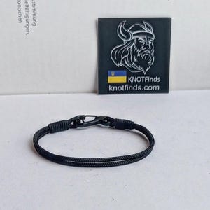 May include: Black paracord bracelet with a black metal clasp. The bracelet is made of two strands of paracord and has a simple design. The bracelet is shown on a white surface.
